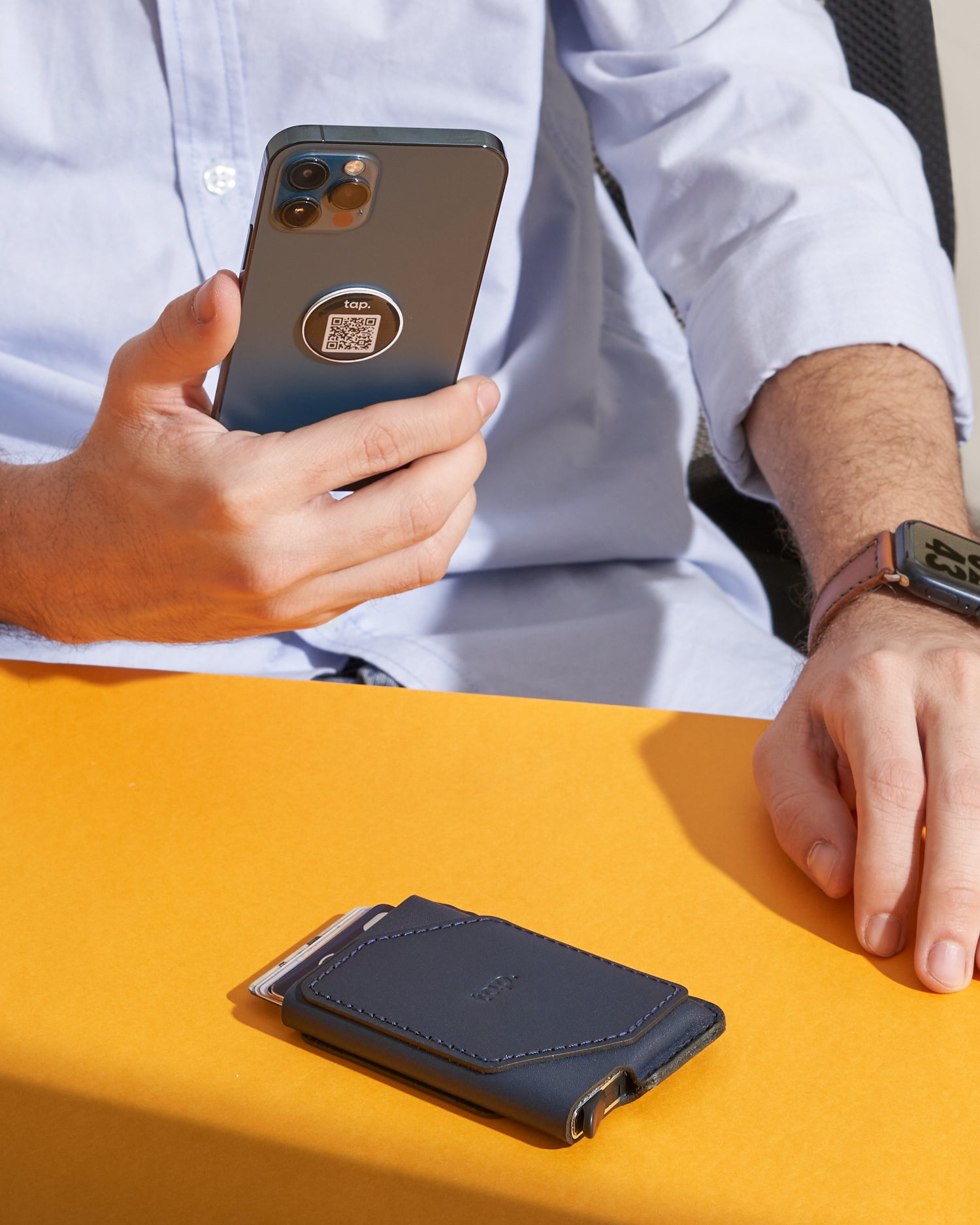 Man holding smartphone with Taps QR code sticker, beside a leather wallet on orange surface.